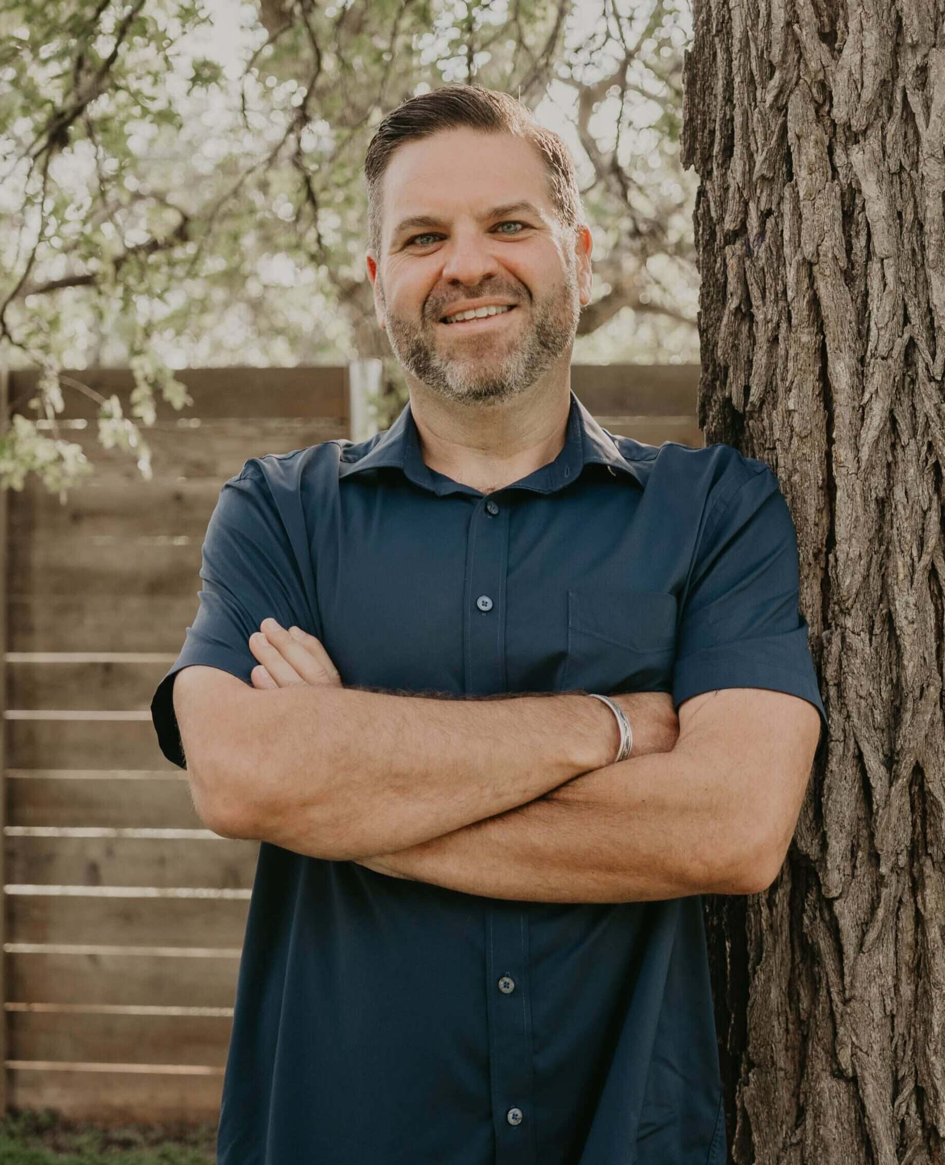 Smiling man standing outdoors near a tree with crossed arms.