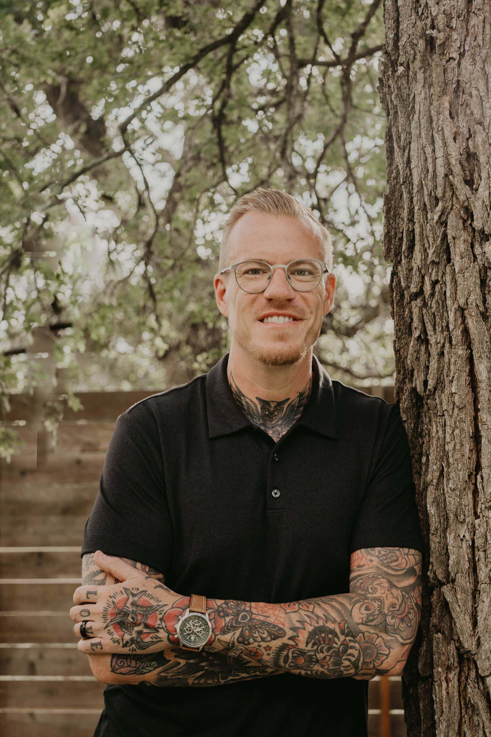 Smiling man with tattoos and glasses standing outdoors near a tree.