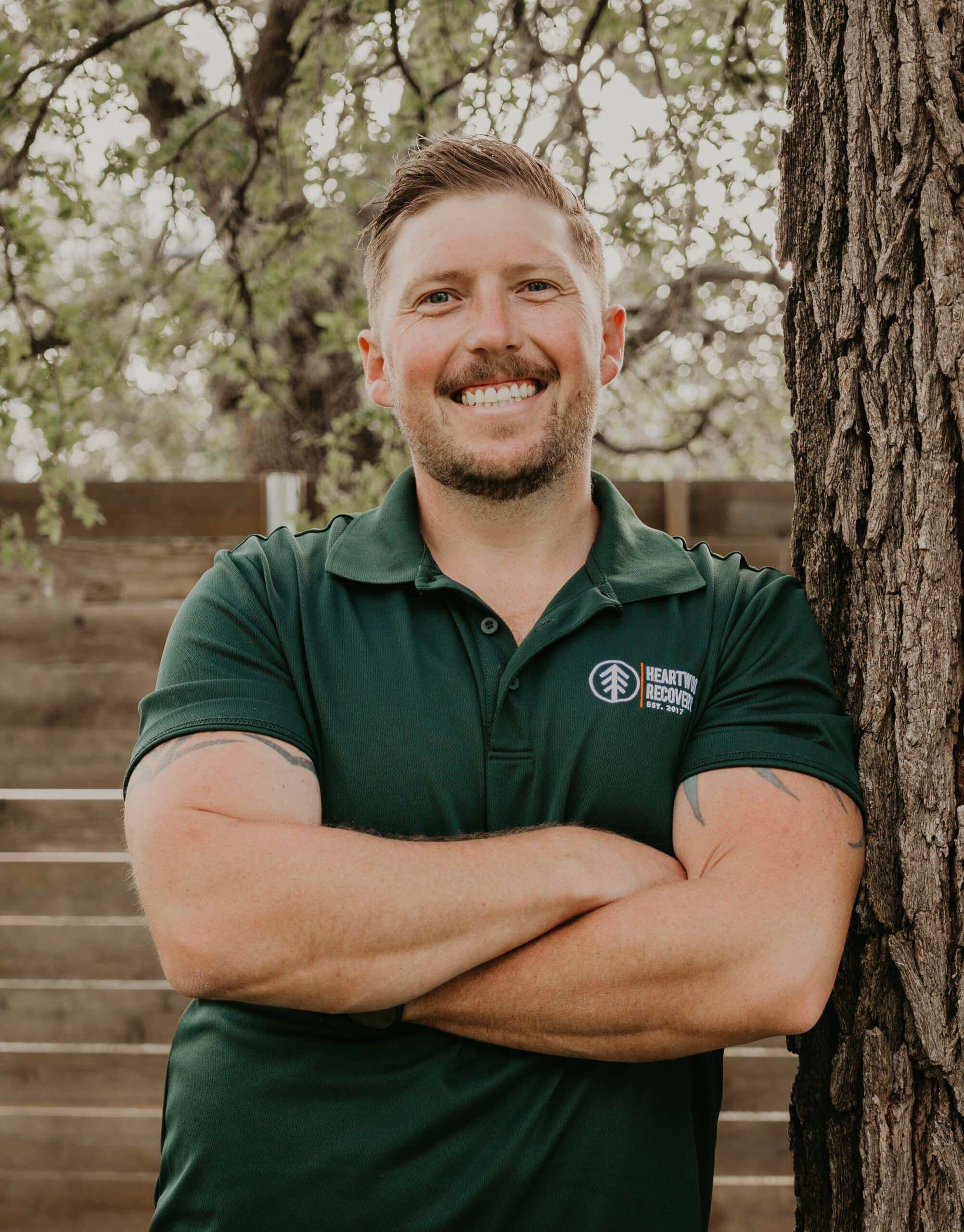 Smiling man in green Heartwood Recovery polo shirt outdoors near a tree.